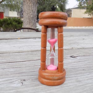 May include: A wooden hourglass featuring a brown frame with rounded top and bottom caps connected by four vertical pillars. The central glass chamber contains bright pink sand, with a portion of the sand currently flowing through the narrow neck.
