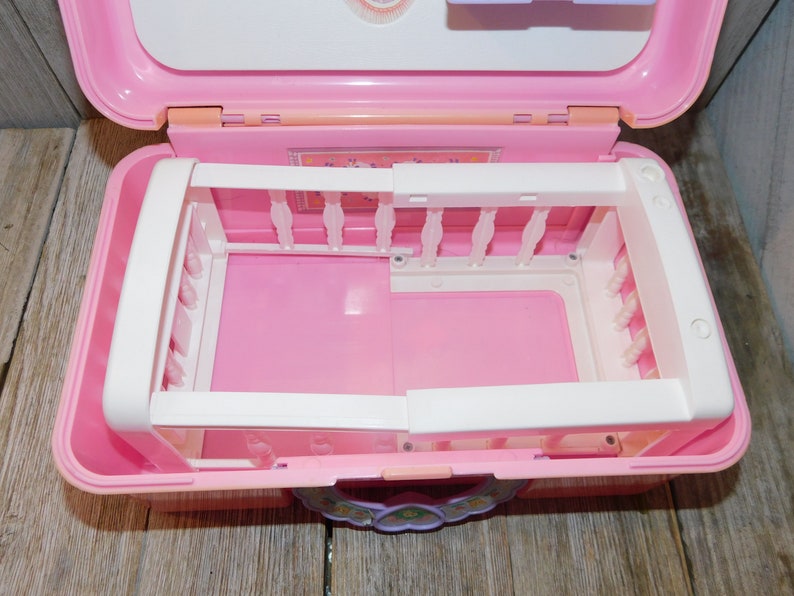 Puede incluir: Cuna de juguete de pl&aacute;stico rosa con barrotes blancos y un colch&oacute;n rosa. La cuna est&aacute; dentro de una maleta de transporte de pl&aacute;stico rosa con un dise&ntilde;o floral.