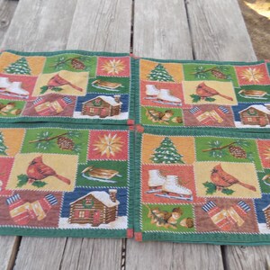 May include: A set of four rectangular placemats featuring a patchwork design of winter scenes. Each placemat displays illustrations of white ice skates, red cardinals, pine cones, evergreen trees, wooden cabins, and striped mittens on a green border.