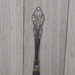 May include: Close-up of a silver butter knife with an ornate handle. The handle features a detailed floral design and is set against a light wood background. The knife is a classic piece of silverware.