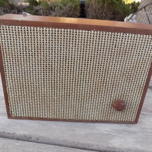 May include: A vintage wooden radio with a woven speaker cover. The radio has a dark brown wooden frame and a light brown woven front. A small, round knob is on the front. The radio is sitting on a weathered wooden surface.