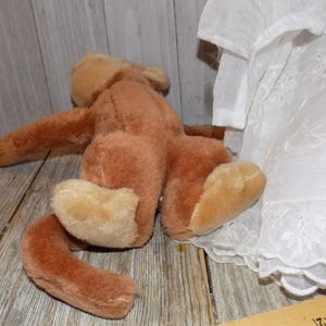 May include: A vintage brown plush toy monkey with tan paws and a long tail, lying on a wooden surface. A white lace-trimmed garment is partially visible on the right side of the image.