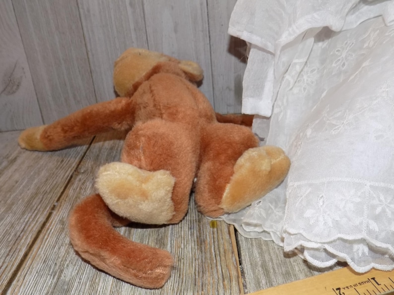 May include: A vintage brown plush toy monkey with tan paws and a long tail, lying on a wooden surface. A white lace-trimmed garment is partially visible on the right side of the image.