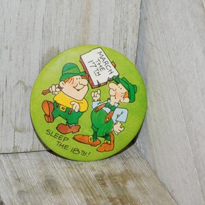 Puede incluir: Un bot&oacute;n verde con dos duendes de dibujos animados. Un duende sostiene un cartel que dice "March the 17th!" El otro duende est&aacute; se&ntilde;alando el cartel. El bot&oacute;n tambi&eacute;n dice "Sleep the 18th!"
