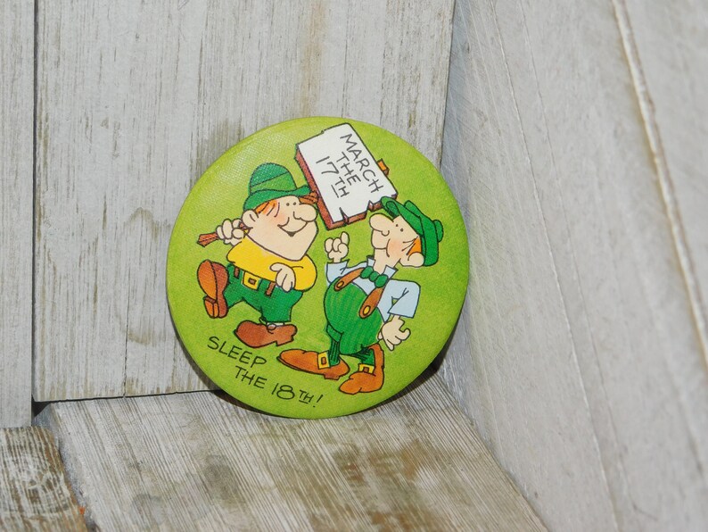 Puede incluir: Un bot&oacute;n verde con dos duendes de dibujos animados. Un duende sostiene un cartel que dice "March the 17th!" El otro duende est&aacute; se&ntilde;alando el cartel. El bot&oacute;n tambi&eacute;n dice "Sleep the 18th!"