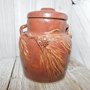 May include: A brown ceramic jar with a lid, featuring handles and a decorative pine branch design. The jar is a warm reddish-brown color, with a textured surface and a rustic aesthetic. The jar is approximately 10 inches tall.