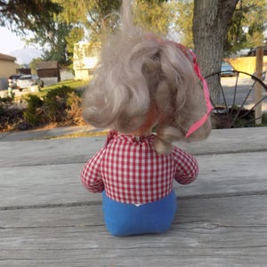 Puede incluir: Una mu&ntilde;eca vintage con cabello rubio que lleva un vestido a cuadros rojos y blancos y pantalones cortos azules. La mu&ntilde;eca est&aacute; sentada sobre una superficie de madera.