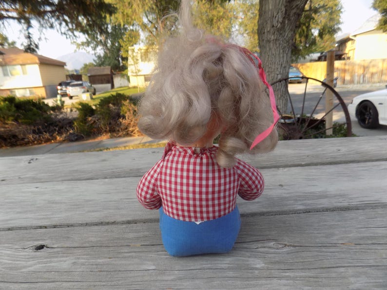 Puede incluir: Una mu&ntilde;eca vintage con cabello rubio que lleva un vestido a cuadros rojos y blancos y pantalones cortos azules. La mu&ntilde;eca est&aacute; sentada sobre una superficie de madera.