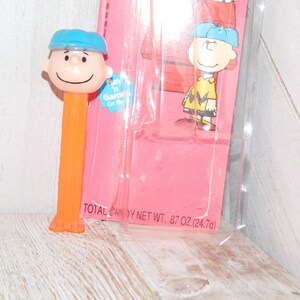 May include: An orange PEZ dispenser featuring a cartoon character with a blue hat and smiling face. The dispenser is in its original packaging, which includes a pink backing card with a cartoon illustration and the text "Fun 'n Games On the Go".