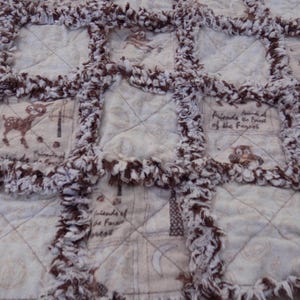 May include: A rag quilt featuring a grid of square fabric patches with frayed brown and white edges. The patches are printed with illustrations of forest animals, trees, and the text Friends of the Forest in a light beige and brown color palette.