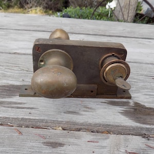 May include: An antique brass door lock set with a key inserted. The set includes a doorknob, a keyhole, and a rectangular base. The metal has a weathered appearance, suggesting age and use. The lock is likely for a vintage door.