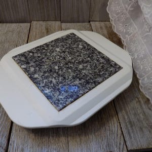May include: A white serving tray with a square, speckled granite insert. The tray has a slightly raised edge and sits on a wooden surface. The granite insert is approximately 6 inches square.