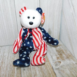 May include: A white Beanie Baby bear wearing a red, white, and blue American flag pattern. The bear has black eyes and a black nose. The bear is sitting on a wooden surface.