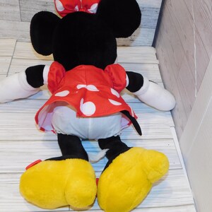 Op de afbeelding: Een pluche Minnie Mouse pop, gezien van achteren, met zwarte oren, een rode jurk met witte stippen en een bijpassende strik. De pop heeft witte handschoenen, zwarte armen en benen en grote gele schoenen.
