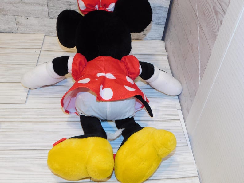 Op de afbeelding: Een pluche Minnie Mouse pop, gezien van achteren, met zwarte oren, een rode jurk met witte stippen en een bijpassende strik. De pop heeft witte handschoenen, zwarte armen en benen en grote gele schoenen.