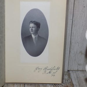 May include: A vintage black and white photograph of a young man in a suit and tie, set within an oval frame. The photograph is mounted on a cream-colored card, with handwritten text including the name "Jay Redfield" and the year '89.