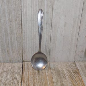 May include: A silver-colored ladle with a rounded bowl and a long handle. The handle has a curved design at the top. The ladle is made of metal and is likely used for serving soups or sauces. The background is a wooden surface.