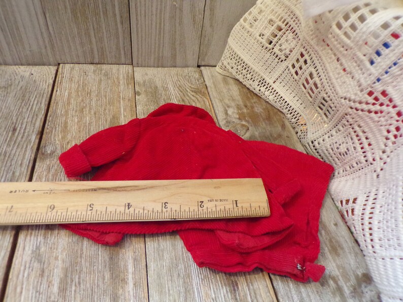 K&ouml;nnte beinhalten: Rote Cord-Samt-Puppenkleidung, ein Pullover und eine Hose, auf einer Holzoberfl&auml;che mit einem Lineal zum Ma&szlig;stab ausgelegt. Der Pullover hat einen Rei&szlig;verschluss.