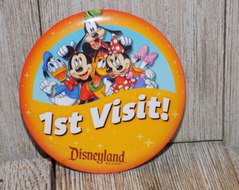 May include: A round, orange Disneyland Resort button with the text "1st Visit!" and images of Mickey Mouse, Minnie Mouse, Donald Duck, Goofy, and Daisy Duck. The button has a light blue background and a yellow border with stars.