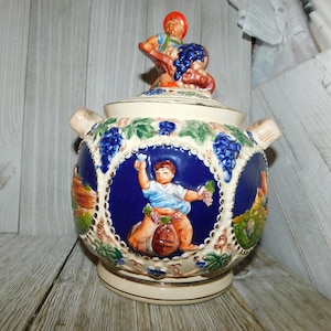 May include: A ceramic pitcher with a lid, decorated with grapevines, a man carrying grapes, and a man holding a glass of wine. The pitcher is painted in shades of blue, green, white, and gold.