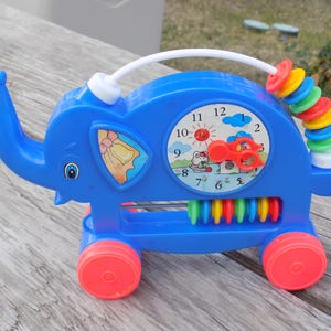 May include: A blue plastic elephant-shaped toy with red wheels and a white handle. The toy features a clock face, an abacus, and a bead maze. The clock shows the time as 1 o'clock. The toy is designed for young children.