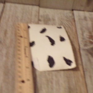 May include: A small, cream-coloured object featuring irregular black spots, reminiscent of a cowhide pattern. A wooden ruler is positioned beside the object. The background is a light-coloured wooden surface.