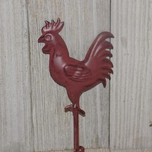 May include: A burgundy metal rooster wall hook. The rooster is in a standing position with a detailed comb, beak, and tail feathers. The hook is attached to a vertical rod with a rounded end. The background is a light wood panel.