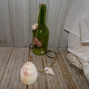 May include: A green glass bottle with a seashell wind chime. The bottle has a jute string and seashell accents. The wind chime has a large seashell and two smaller shells. The background is a wooden surface.