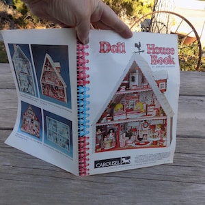 May include: A vintage book titled "Doll House Book" by Jean Rae Thornton. The book features a colorful illustration of a dollhouse with red and white accents. The book is open to a page showing a detailed interior view of the dollhouse.