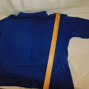 May include: A royal blue long-sleeved turtleneck shirt with a mock neck collar. The shirt is laid flat on a white surface with a wooden ruler next to it.