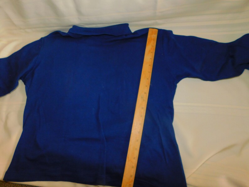 May include: A royal blue long-sleeved turtleneck shirt with a mock neck collar. The shirt is laid flat on a white surface with a wooden ruler next to it.