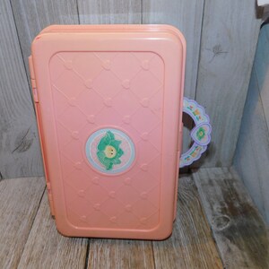 Puede incluir: Una casa de juegos de pl&aacute;stico rosa con un asa en forma de coraz&oacute;n y un dise&ntilde;o floral. La casa de juegos tiene un dise&ntilde;o de flor blanca y verde en la parte delantera.