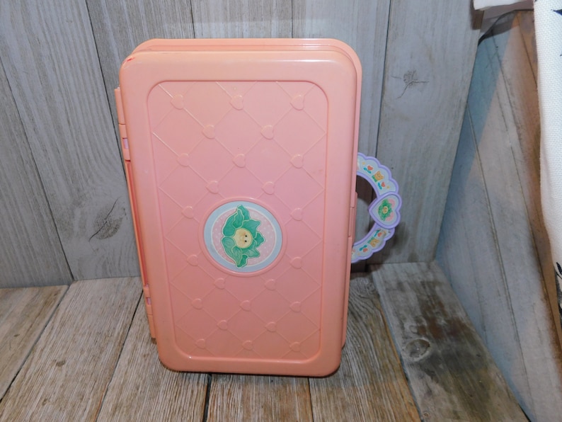 Puede incluir: Una casa de juegos de pl&aacute;stico rosa con un asa en forma de coraz&oacute;n y un dise&ntilde;o floral. La casa de juegos tiene un dise&ntilde;o de flor blanca y verde en la parte delantera.