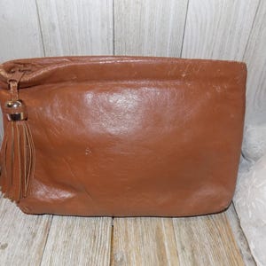 May include: A brown leather clutch with a tassel. The clutch has a zipper closure and a smooth leather finish.