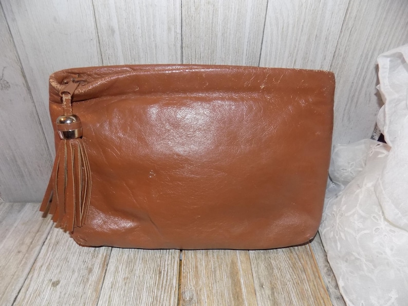 May include: A brown leather clutch with a tassel. The clutch has a zipper closure and a smooth leather finish.