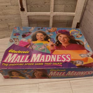 May include: A rectangular board game box for Electronic Mall Madness, the shopping spree game that talks. The box features purple and blue graphics with images of two people, shopping bags, and play money. The title is printed in large pink text.