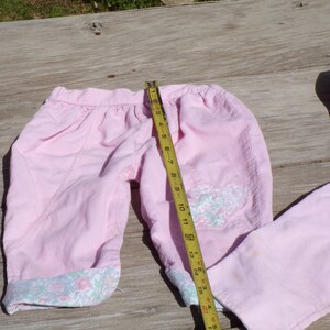 Puede incluir: Un pantal&oacute;n de pana rosa con ribete floral y una aplicaci&oacute;n de mariposa. El pantal&oacute;n est&aacute; colocado sobre una superficie de madera con una cinta m&eacute;trica.