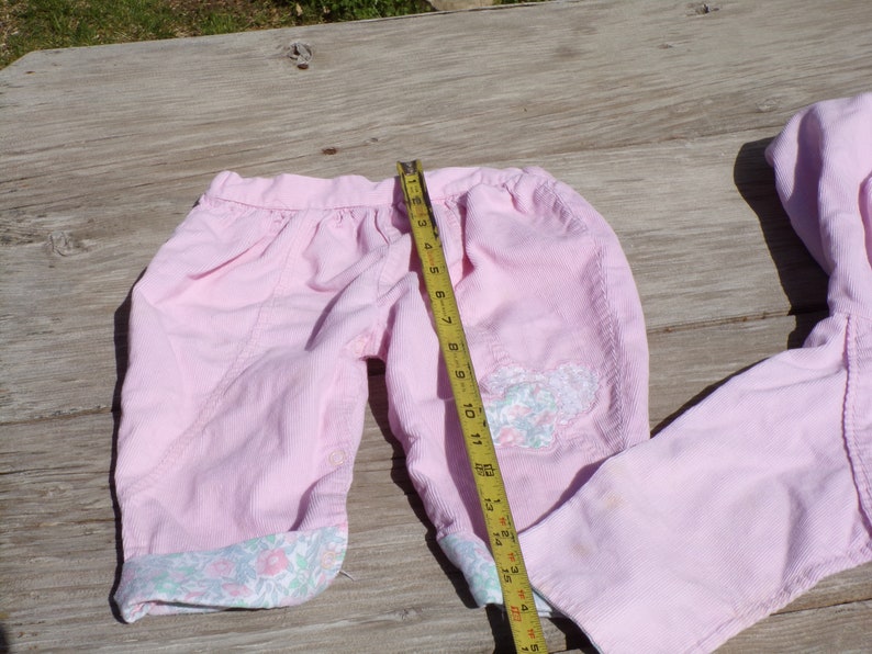Puede incluir: Un pantal&oacute;n de pana rosa con ribete floral y una aplicaci&oacute;n de mariposa. El pantal&oacute;n est&aacute; colocado sobre una superficie de madera con una cinta m&eacute;trica.