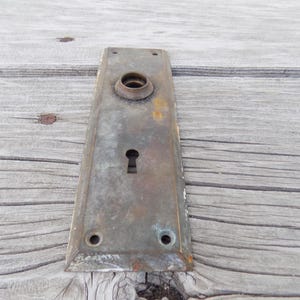 May include: A rectangular, weathered brass door plate featuring a central circular knob housing and a keyhole below. The metal surface displays a tarnished, aged patina with visible oxidation, four small screw holes, and beveled edges.