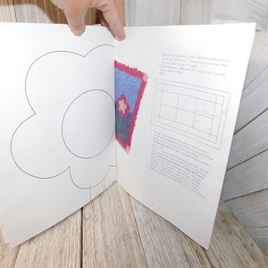 May include: A book with a white background and a black outline of a flower. The book is open to a page with instructions on how to make a rug. The instructions include a grid with squares and a picture of a blue and pink rug with a flower design.