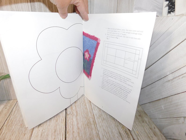 May include: A book with a white background and a black outline of a flower. The book is open to a page with instructions on how to make a rug. The instructions include a grid with squares and a picture of a blue and pink rug with a flower design.