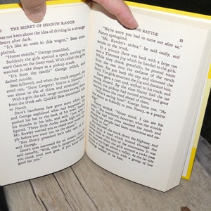 May include: Open book with yellow pages showing a scene from a novel about a ranch. The text on the page reads "The Secret of Shadow Ranch" and "We're sorry you had to come out after us."