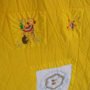 May include: Close-up of a bright yellow quilt with embroidered patches. The patches feature colorful designs, including a rainbow, a bear, and text. The quilt has a quilted diamond pattern throughout.