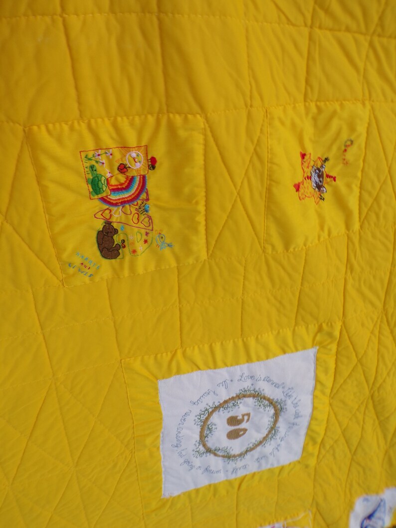 May include: Close-up of a bright yellow quilt with embroidered patches. The patches feature colorful designs, including a rainbow, a bear, and text. The quilt has a quilted diamond pattern throughout.