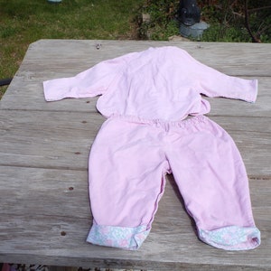 Puede incluir: Un conjunto de pijama rosa de dos piezas con una parte superior de manga larga y pantalones. Los pantalones tienen un ribete floral alrededor de los pu&ntilde;os.