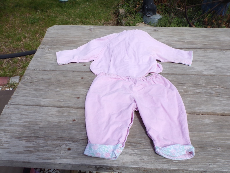 Puede incluir: Un conjunto de pijama rosa de dos piezas con una parte superior de manga larga y pantalones. Los pantalones tienen un ribete floral alrededor de los pu&ntilde;os.