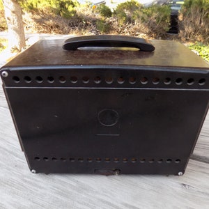 Puede incluir: Una caja vintage de color marr&oacute;n oscuro con un asa negra en la parte superior. La caja tiene una serie de peque&ntilde;os agujeros espaciados uniformemente a lo largo de los bordes superior e inferior. Un emblema circular est&aacute; centrado en la parte frontal.