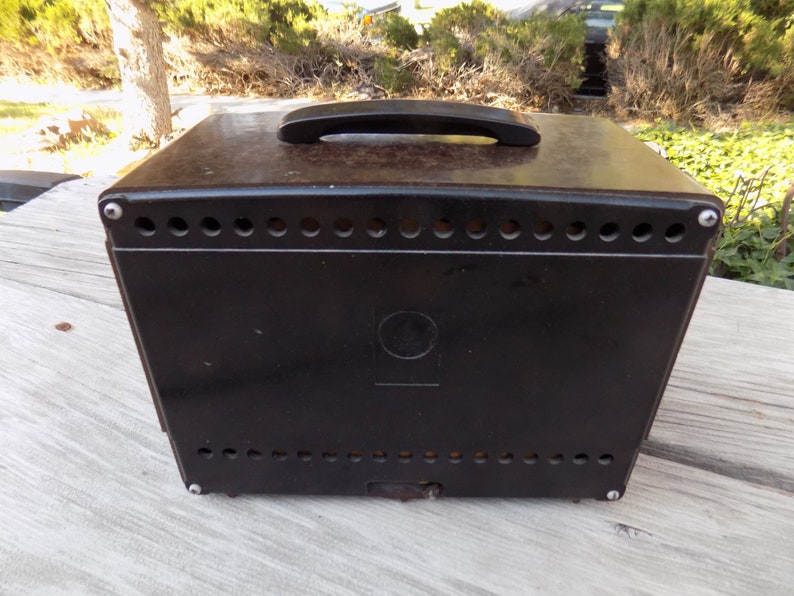 Puede incluir: Una caja vintage de color marr&oacute;n oscuro con un asa negra en la parte superior. La caja tiene una serie de peque&ntilde;os agujeros espaciados uniformemente a lo largo de los bordes superior e inferior. Un emblema circular est&aacute; centrado en la parte frontal.