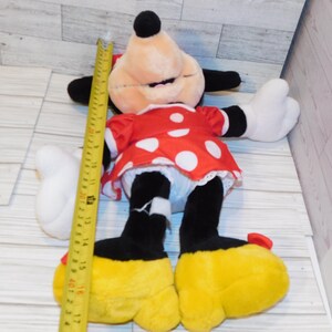 Op de afbeelding: Een Minnie Mouse pluche pop, met een rode jurk met witte stippen, zwarte oren en gele schoenen. De pop is ongeveer 43 cm hoog, met een gele meetlint ernaast.