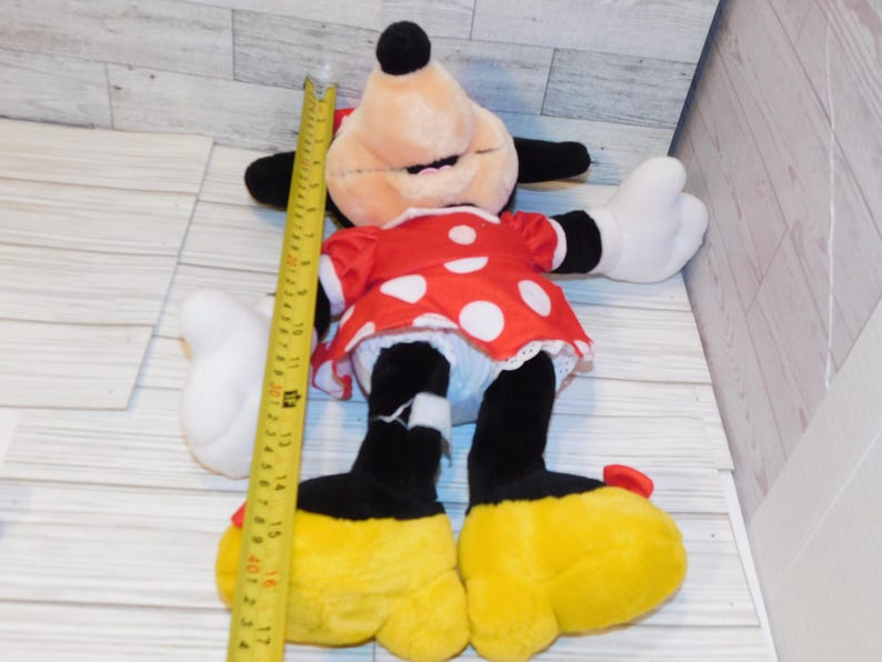 Op de afbeelding: Een Minnie Mouse pluche pop, met een rode jurk met witte stippen, zwarte oren en gele schoenen. De pop is ongeveer 43 cm hoog, met een gele meetlint ernaast.